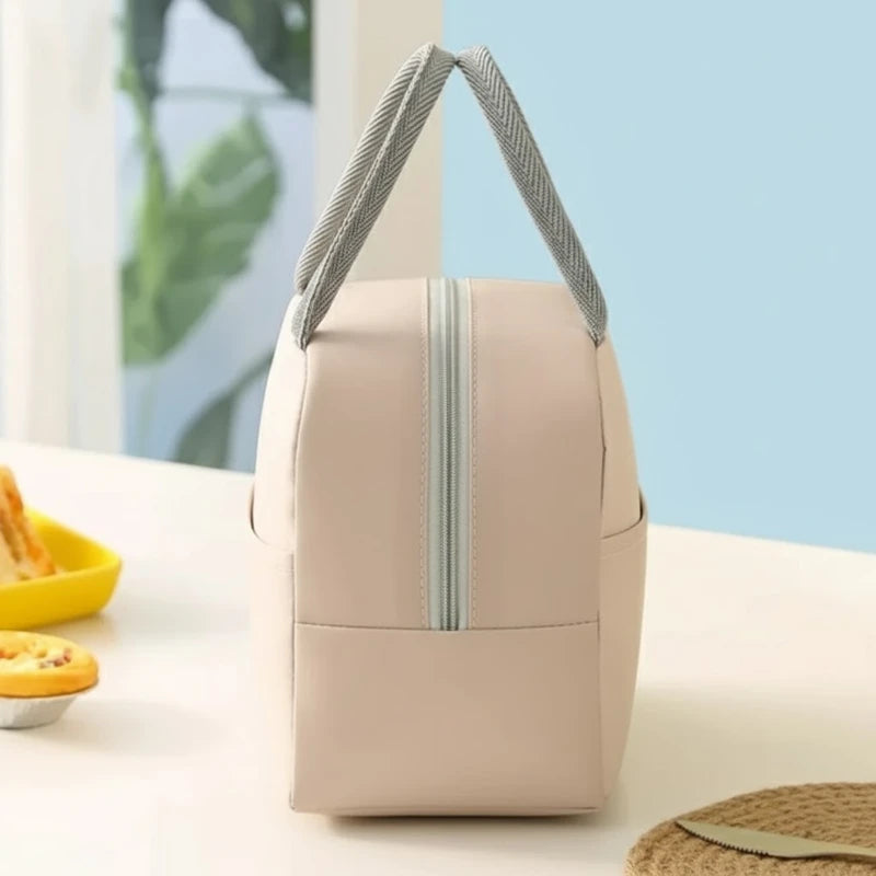 Sac isotherme repas femme beige sans motif, vu de côté, posé sur une table claire, avec des poignées grises et une fermeture zippée verticale, dans un décor lumineux.