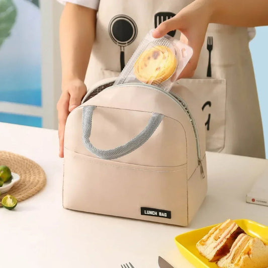 Sac isotherme repas femme beige sans motif, vu de face et ouvert sur le dessus, une main y glisse une tartelette. Le sac est posé sur une table claire avec des sandwichs et des assiettes autour.