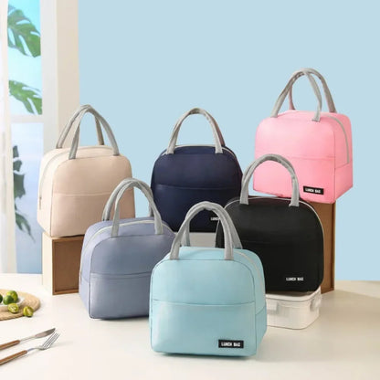 Six sacs isothermes repas avec poignées grises sont disposés sur une table et des supports : on y voit les coloris bleu clair, bleu foncé, rose, beige, noir et gris. En arrière-plan, quelques citrons verts et couverts décorent la scène lumineuse.