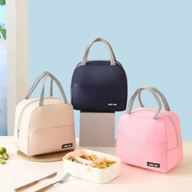 Trois sacs isothermes repas avec poignées grises sont disposés sur une table : un rose, un beige et un bleu marine. Devant eux, une boîte repas ouverte contient un déjeuner appétissant, et quelques citrons verts sont visibles sur le côté.