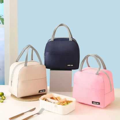 Trois sacs isothermes repas avec poignées grises sont disposés sur une table : un rose, un beige et un bleu marine. Devant eux, une boîte repas ouverte contient un déjeuner appétissant, et quelques citrons verts sont visibles sur le côté.