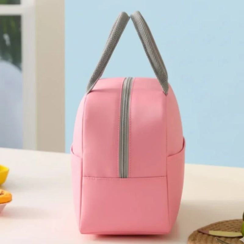 Vue latérale d’un sac isotherme rose clair avec poignées grises, posé sur une table blanche. En arrière-plan, on distingue une assiette jaune et quelques citrons verts sur un dessous de verre tressé.