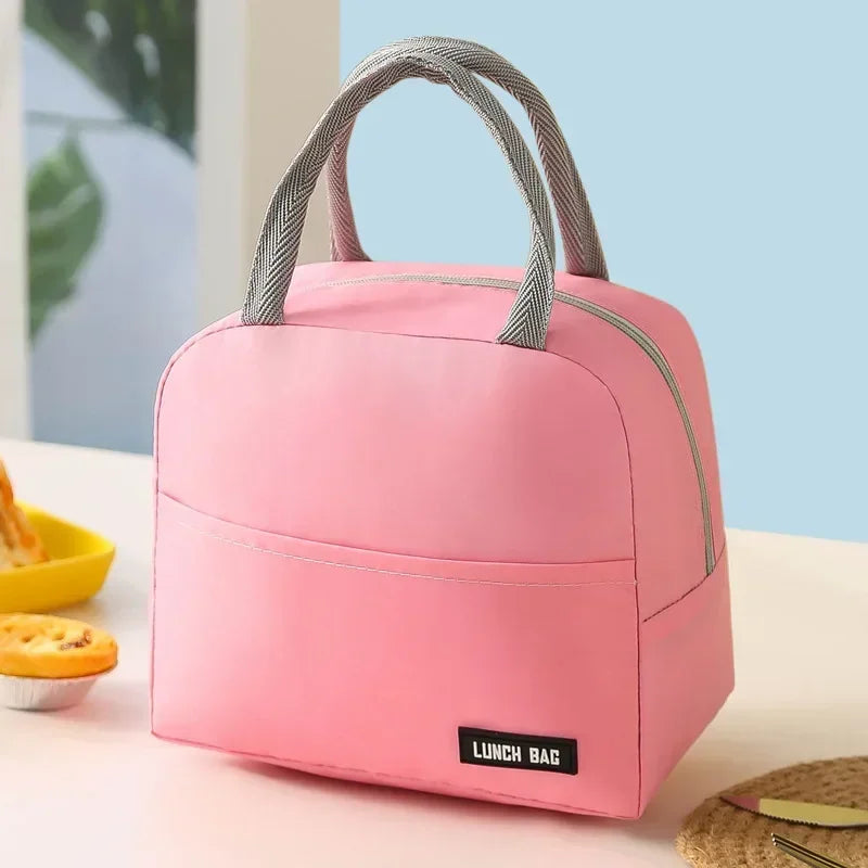 Sac isotherme repas rose posé sur une table claire, avec deux petites tartes sur le côté et une assiette jaune à l’arrière-plan.