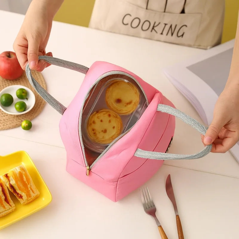 Vue de dessus d’un sac isotherme rose ouvert contenant deux petits gâteaux dans un récipient transparent, posé sur une table avec des couverts, une assiette de sandwichs et quelques citrons verts.