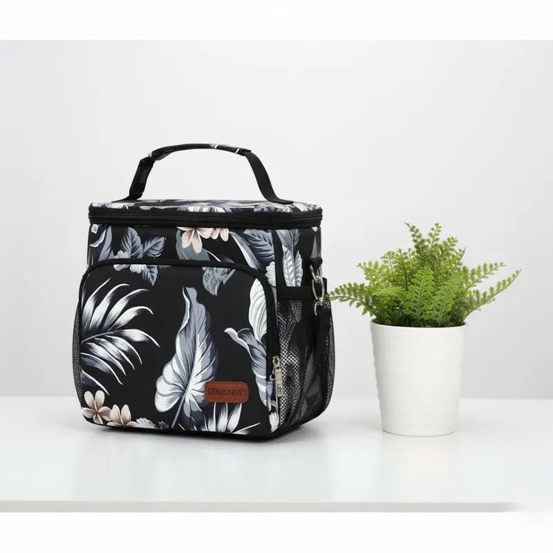 Sac isotherme repas noir à motif floral posé sur une table blanche, idéal pour transporter un déjeuner frais.