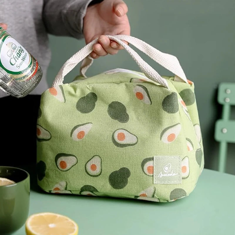 Sac isotherme vert clair décoré de motifs d’avocats, tenu par une main au-dessus d’une table verte avec un demi-citron et une boisson à côté.