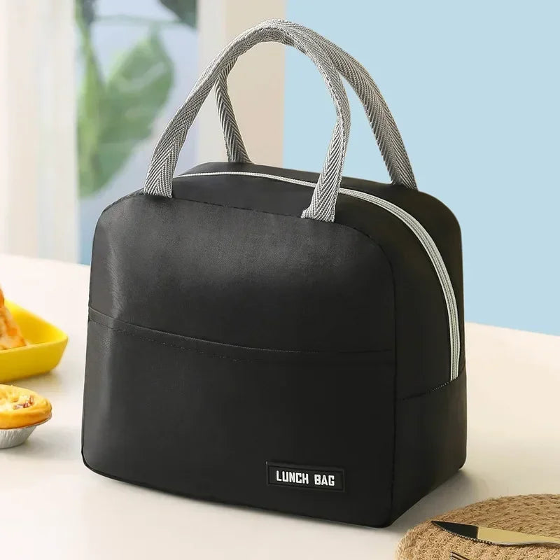 Sac isotherme noir vu de face, posé sur une table blanche, avec poignées grises et fermeture éclair argentée, fond bleu clair et lumière naturelle.