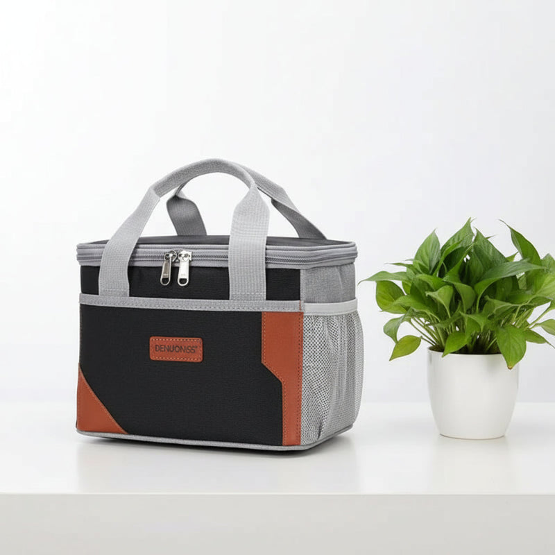 Sac isotherme repas noir et gris avec détails en simili-cuir, posé sur une table blanche à côté d’une plante verte.