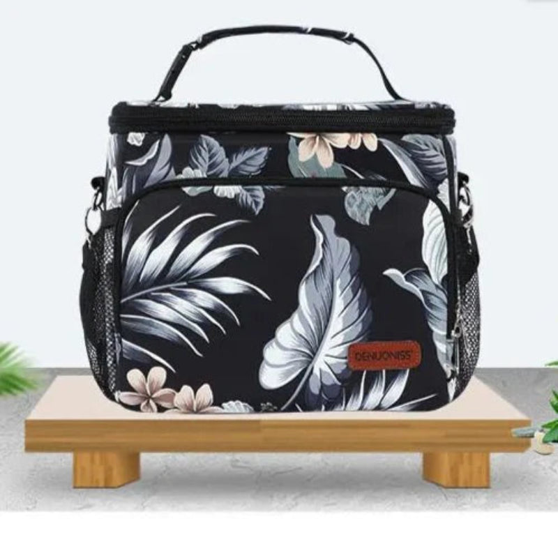 Sac isotherme repas noir à motif floral posé sur un support en bois, avec poche avant et poignée renforcée.