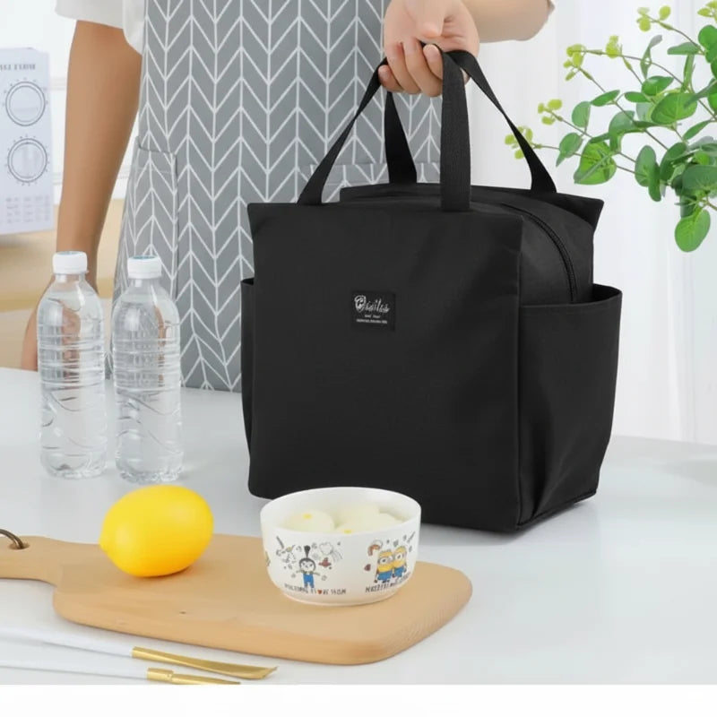 Sac isotherme repas noir type lunch bag porté à la main, avec deux bouteilles d’eau et un bol sur table, idéal pour déjeuner au bureau.