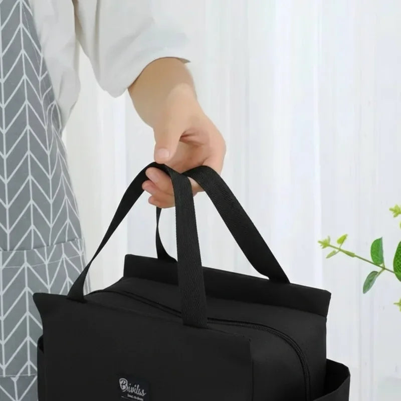 Main tenant un sac isotherme repas noir type lunch bag avec poignées et fermeture zippée, idéal pour transporter un déjeuner au bureau.