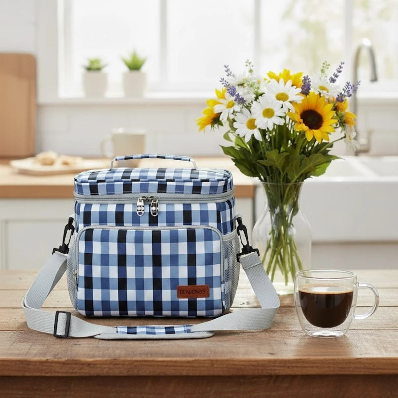 Sac isotherme motif vichy bleu posé sur une table en bois dans une cuisine, avec bandoulière grise, bouquet de fleurs et tasse de café à côté