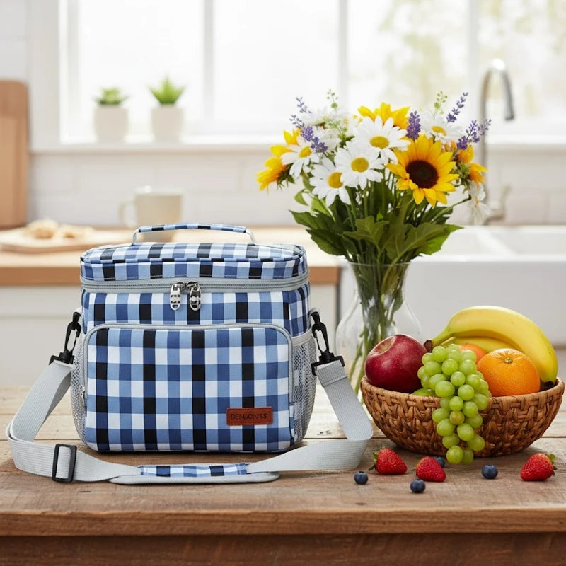 Sac isotherme motif vichy bleu posé sur une table en bois avec une corbeille de fruits, bouquet de fleurs et cuisine en arrière-plan