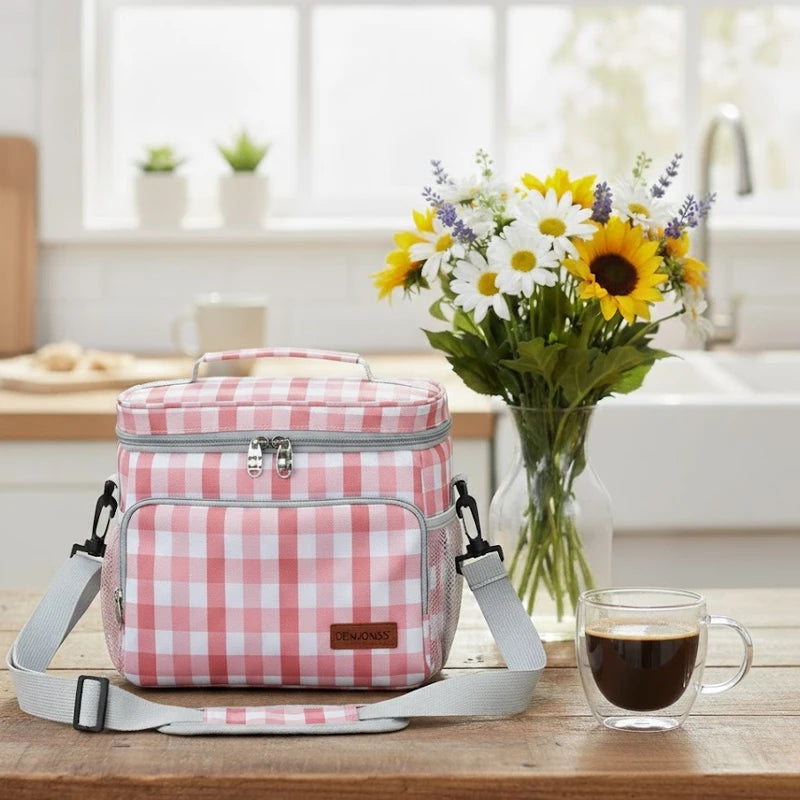 Sac isotherme motif vichy rose posé sur une table en bois dans une cuisine, avec bandoulière grise, bouquet de fleurs et tasse de café en arrière-plan