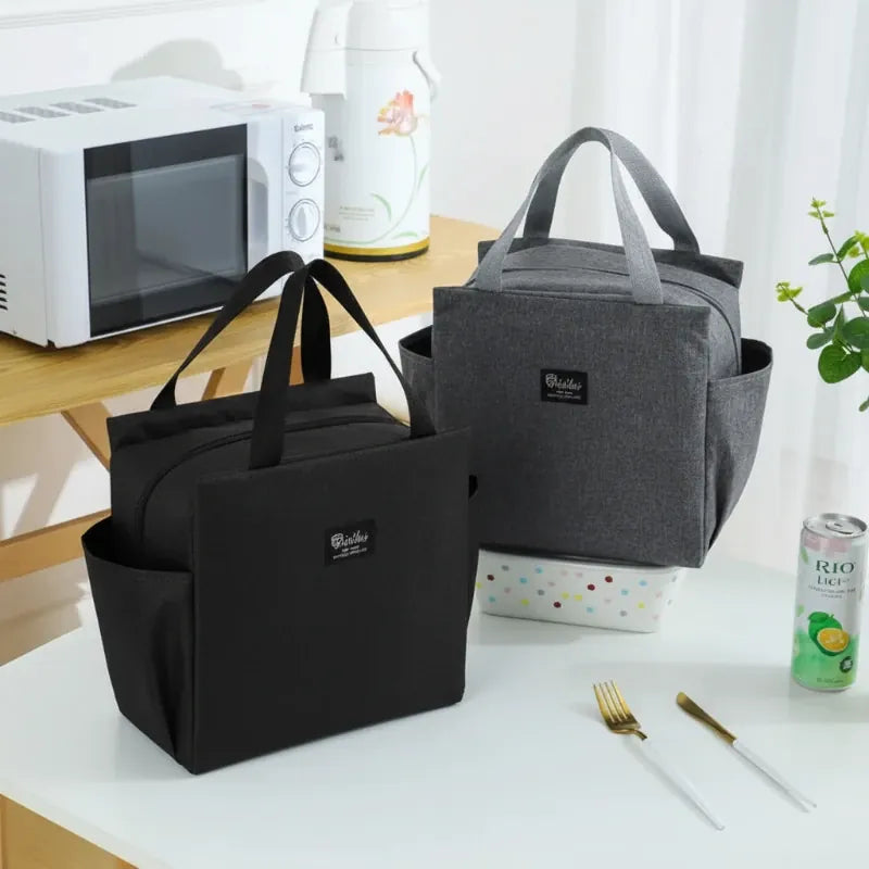 Deux sacs isothermes type lunch bag noir et gris posés sur une table, avec lunch box, boisson et couverts, pour déjeuner au bureau.